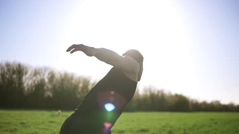 a man in a black t-shirt is doing sports on a green meadow. raises the weight with one hand and holds it on top. backside view