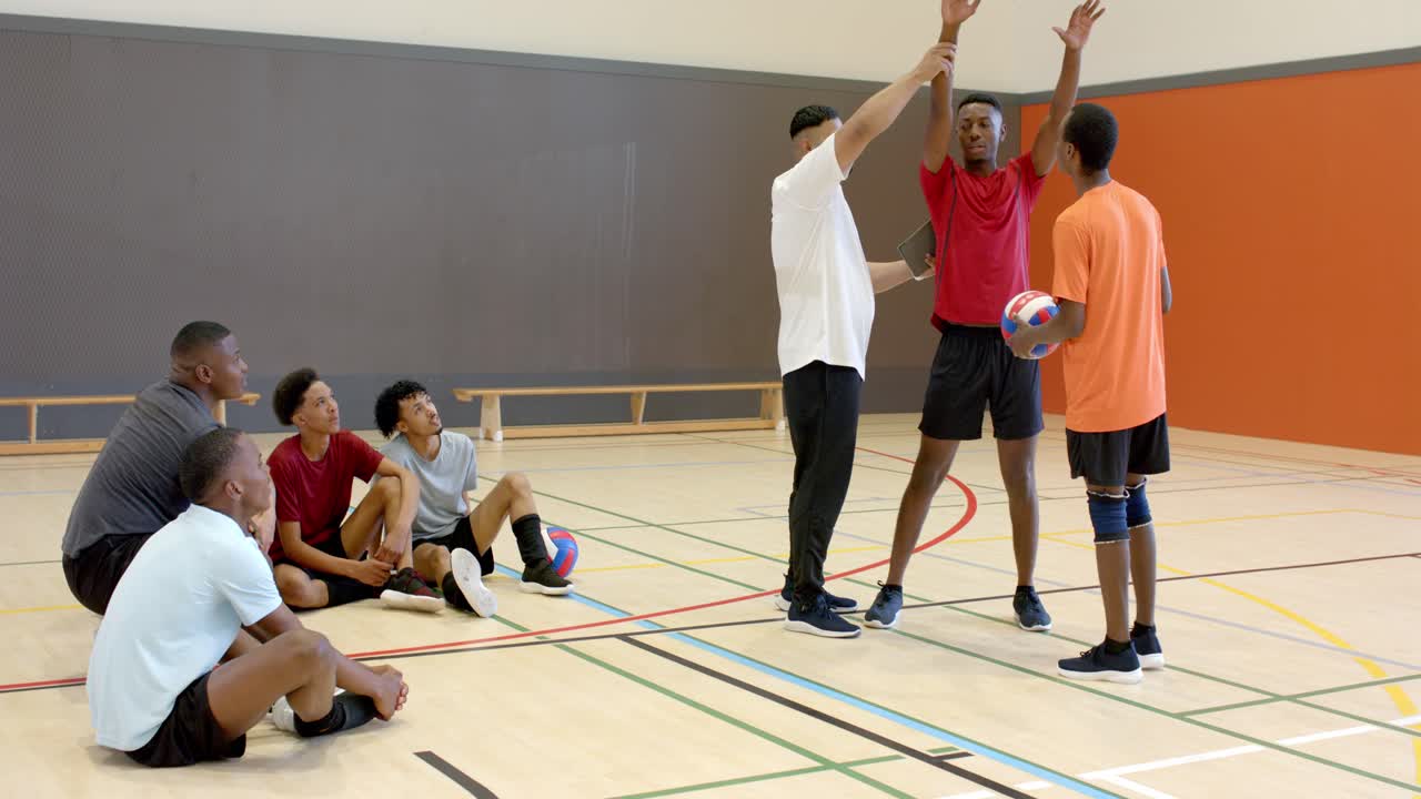 Trainer konsultiert Tablet und leitet roten Spieler beim Blocken, während ein Teamkollege einen Volleyball in der Sporthalle hält.