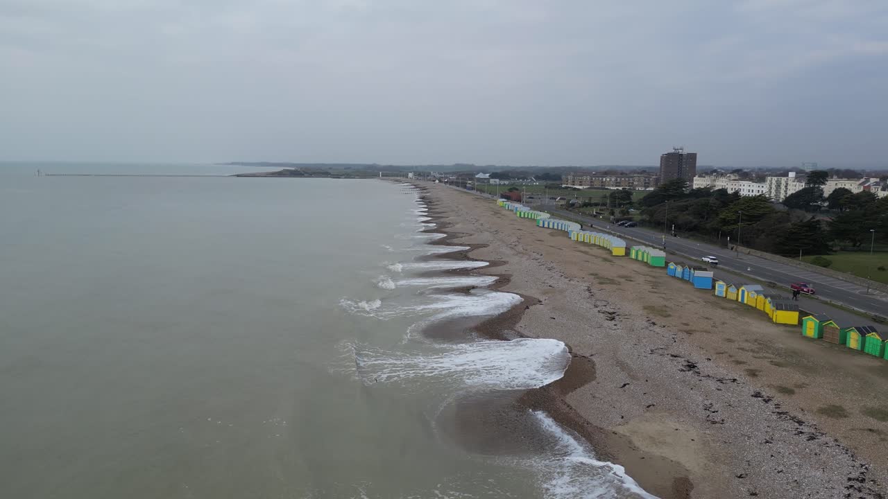 Overcast day on a UK pebble beach. Gentle waves wash the textured shore. Colorful beach huts line the beach next to a road with cars.