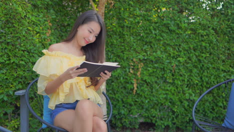 Young-brunette-woman-reading-an-interesting-book-or-a-novel-and-enjoying-the-plot-of-the-story-while-sitting-on-a-chair-in-the-garden