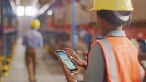 trabajadora afroamericana con casco usando un teléfono inteligente en un almacén