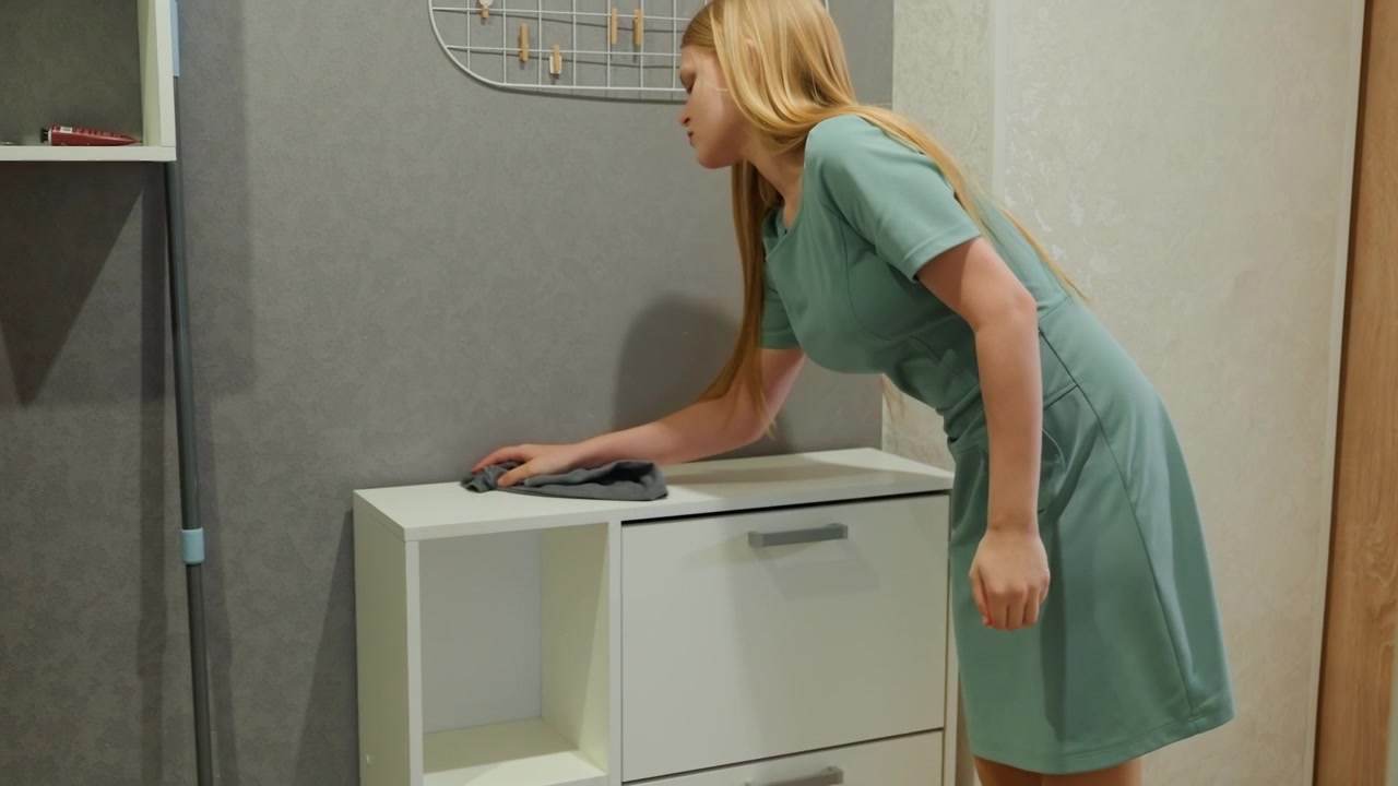 Woman organizing shoe cabinet in entryway, kneeling to adjust drawers and wipe surface with cloth, minimalist white furniture, dusting and arranging storage, tidy foyer and household organization