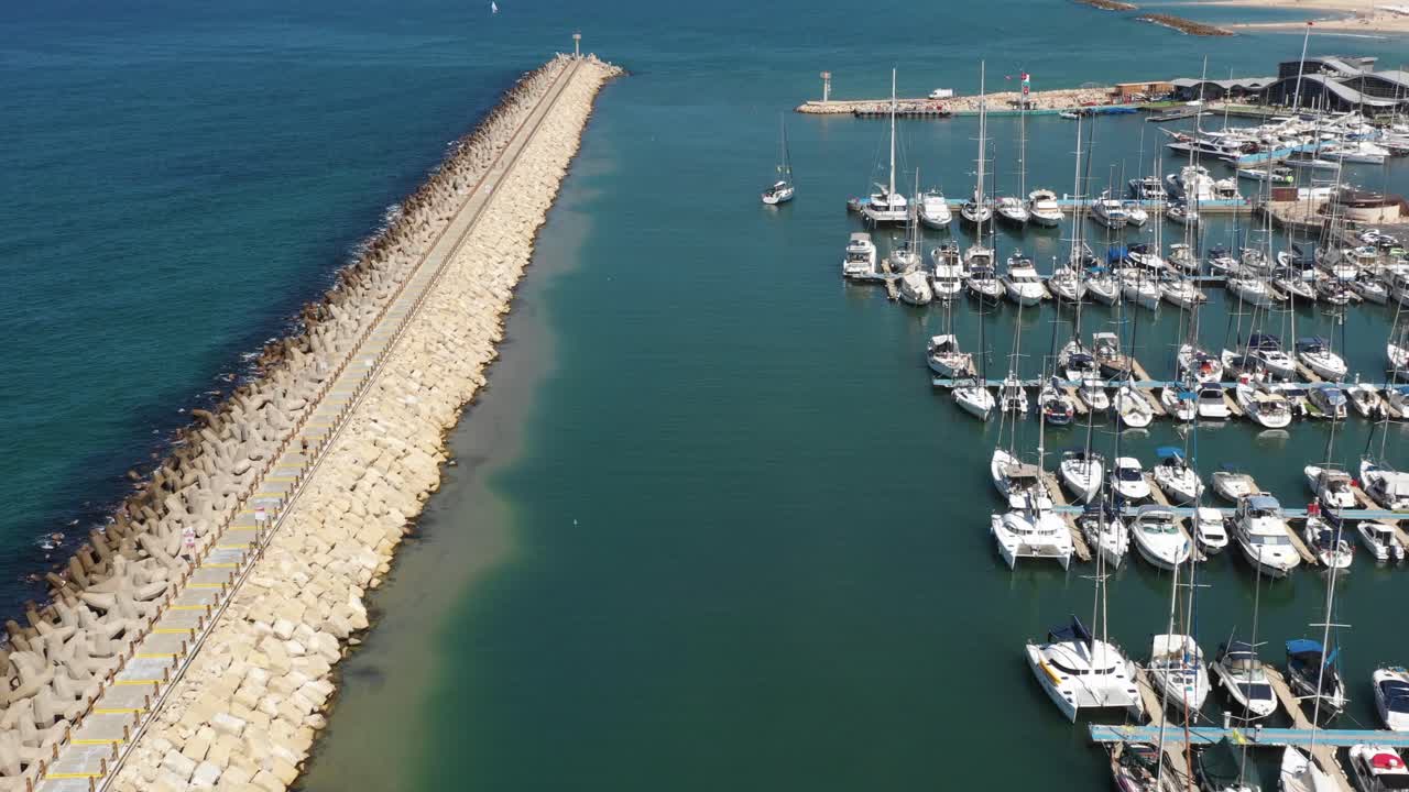 Vários iates à vela navegam calmamente pela marina de Herzeliya, enquanto pequenas ondas quebram ao fundo, em um dia ensolarado