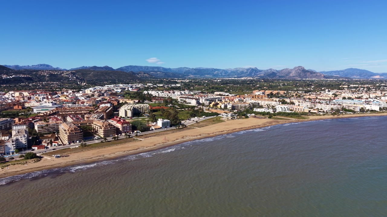 Città costiera di Denia sul Mar Mediterraneo a Valencia, Spagna, che mostra spiaggia sabbiosa, edifici urbani, vegetazione lussureggiante e catena montuosa sotto un cielo azzurro limpido.