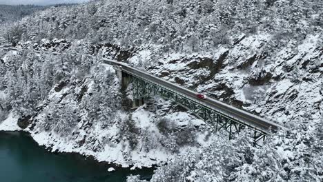 Disparo-De-Un-Dron-De-Un-Coche-Rojo-Que-Cruza-Un-Puente-Elevado-Con-Nieve-Por-Todas-Partes