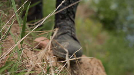 warrior climbs steep dirt slope gripping earth with gloved hand, surrounded by green grass and scattered stones, crawling forward during outdoor mission with head bowed and muscles flexed in effort