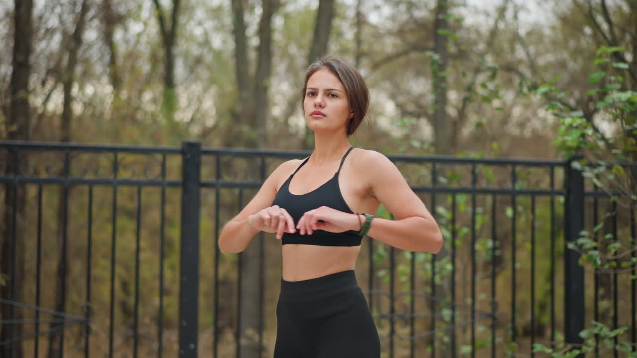 Lady performing fitness exercise outdoors in front of iron fence with trees in background, she is stretching her arms while maintaining a healthy and active lifestyle in natural environment