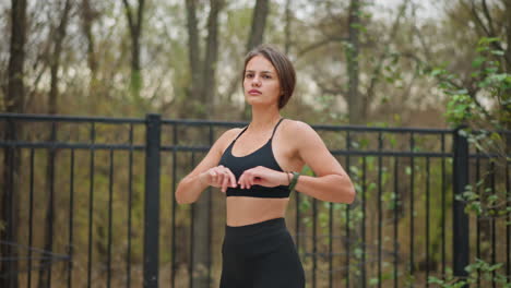 lady performing fitness exercise outdoors in front of iron fence with trees in background, she is stretching her arms while maintaining a healthy and active lifestyle in natural environment
