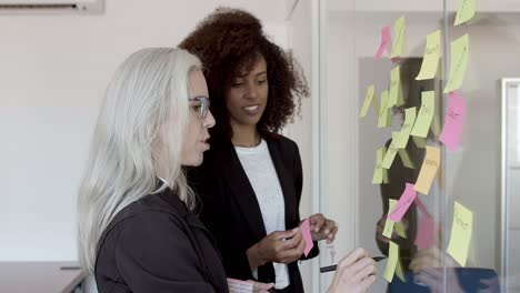 Happy female colleagues sticking memos on glass board