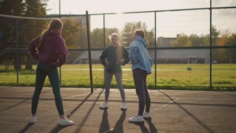 mujeres jóvenes en leggings participan en actividades de fitness en una cancha al aire libre con un campo de fútbol en el fondo y la luz del sol proyectando largas sombras, enfatizando el trabajo en equipo, el ejercicio y un estilo de vida activo