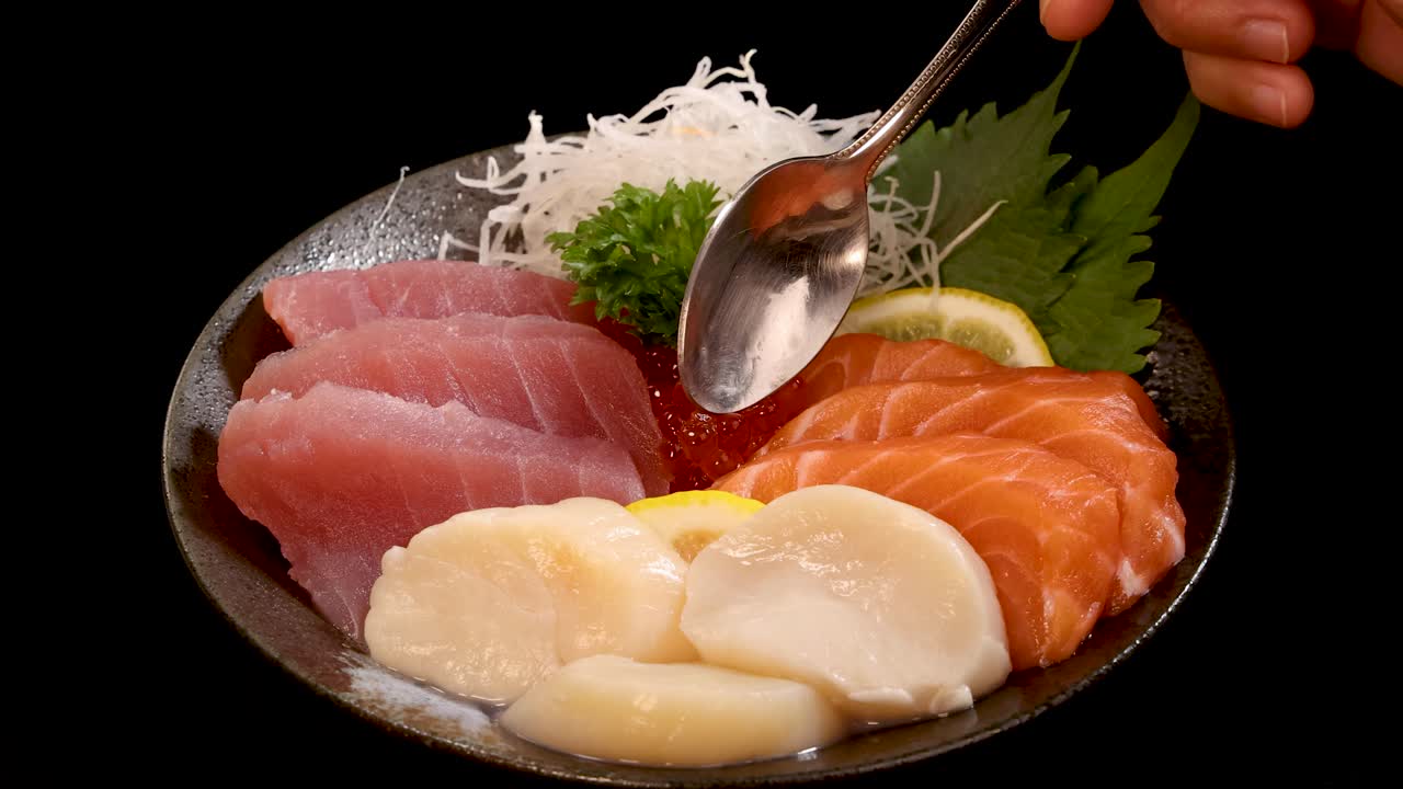 A hand uses a spoon to serve salmon roe onto a plate of assorted sashimi and sushi, arranged on a black background with soft, focused lighting