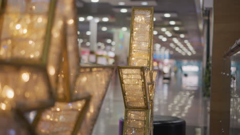 close-up of intricate gold frames with detailed patterns illuminated by warm glowing light, surrounded by blinking white lights, background shows blurred interior of a mall with reflective floors