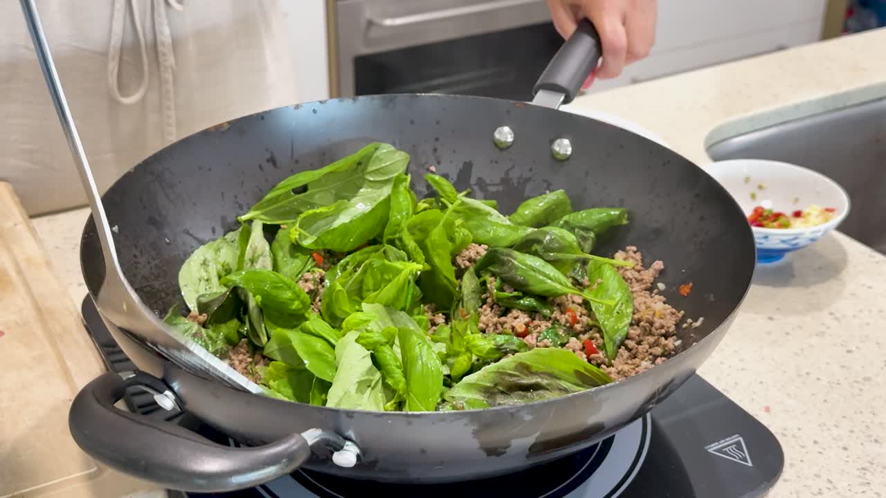 A person stir fries pork and fresh basil leaves in a black wok on an induction cooktop, using a spatula under bright kitchen lighting