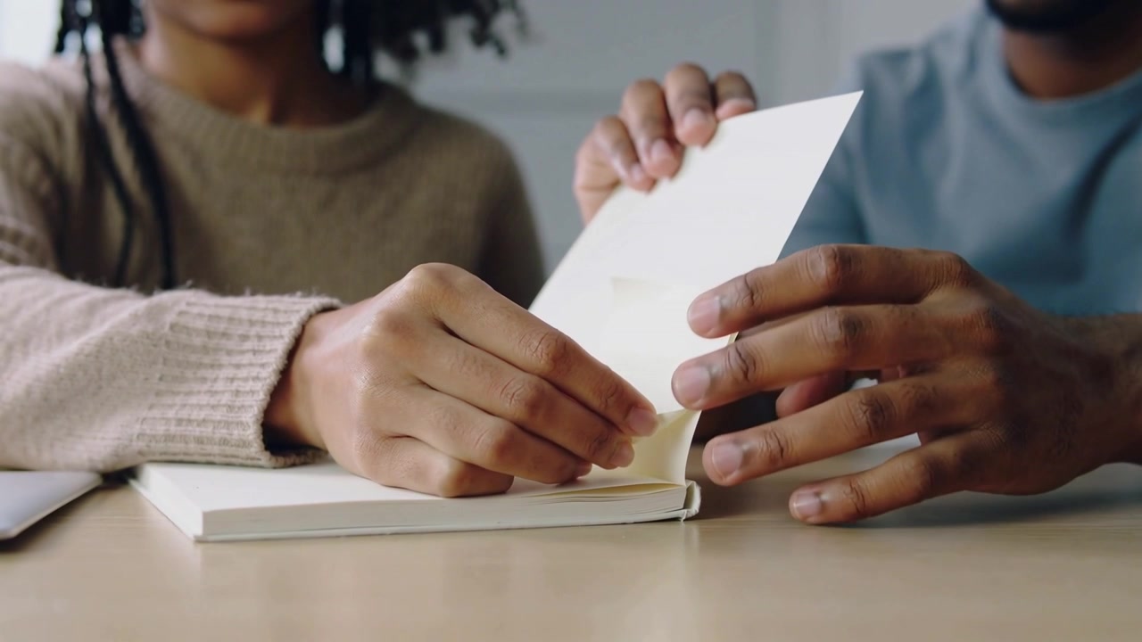 Sollevando la linguetta, una persona con maglione beige e maglietta blu sfoglia pagine e allinea un quaderno sulla scrivania di casa.