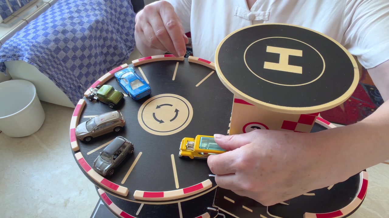 Nærbillede af en anonym kvinde, der bruger sine hænder til at parkere miniature legetøjsbiler i en trælegegarage. Statisk nærbillede set fra oven.