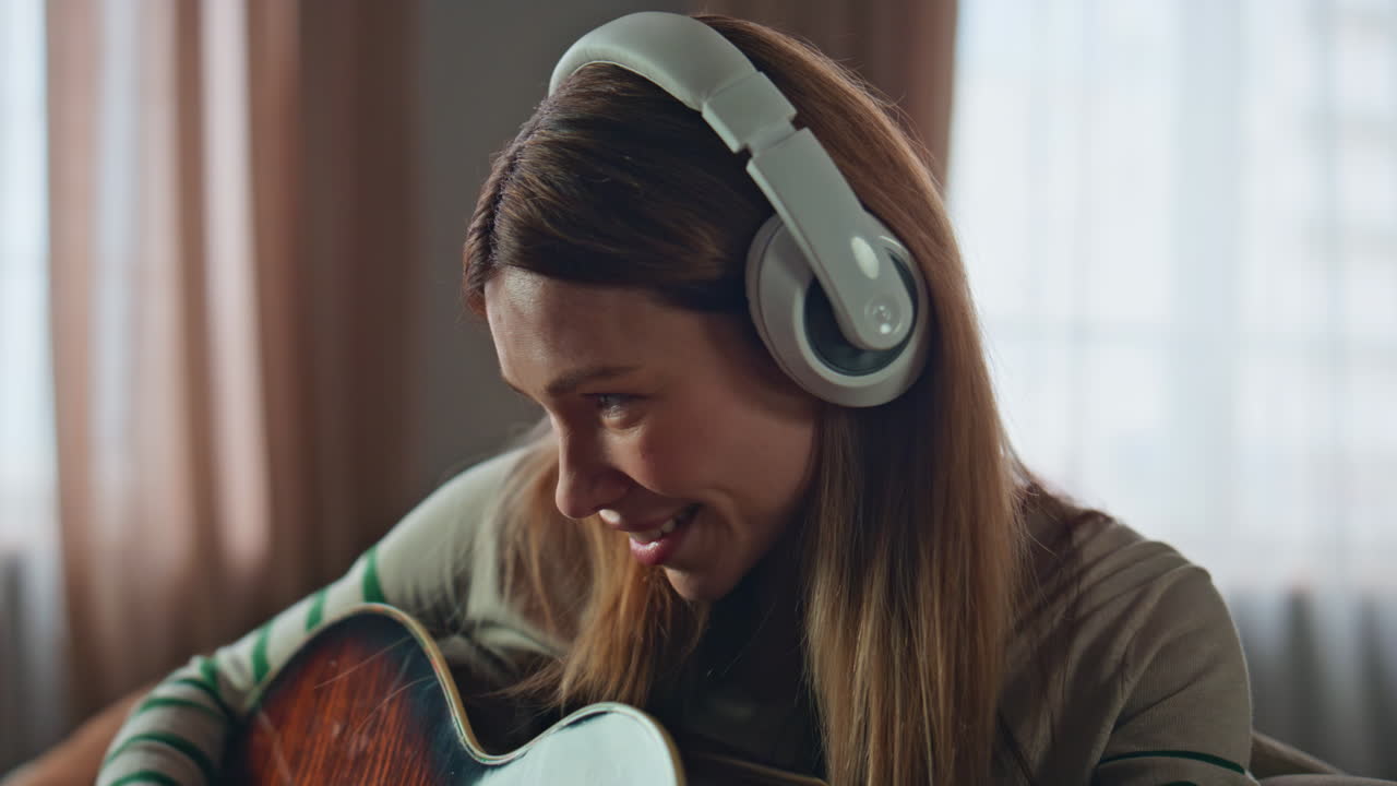 Happy woman practicing guitar playing in earphones at apartment close up.