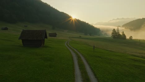 A-woman-walks-on-a-country-road-at-sunrise-in-Wagenbrüchsee,-Germany,-surrounded-by-misty-mountains