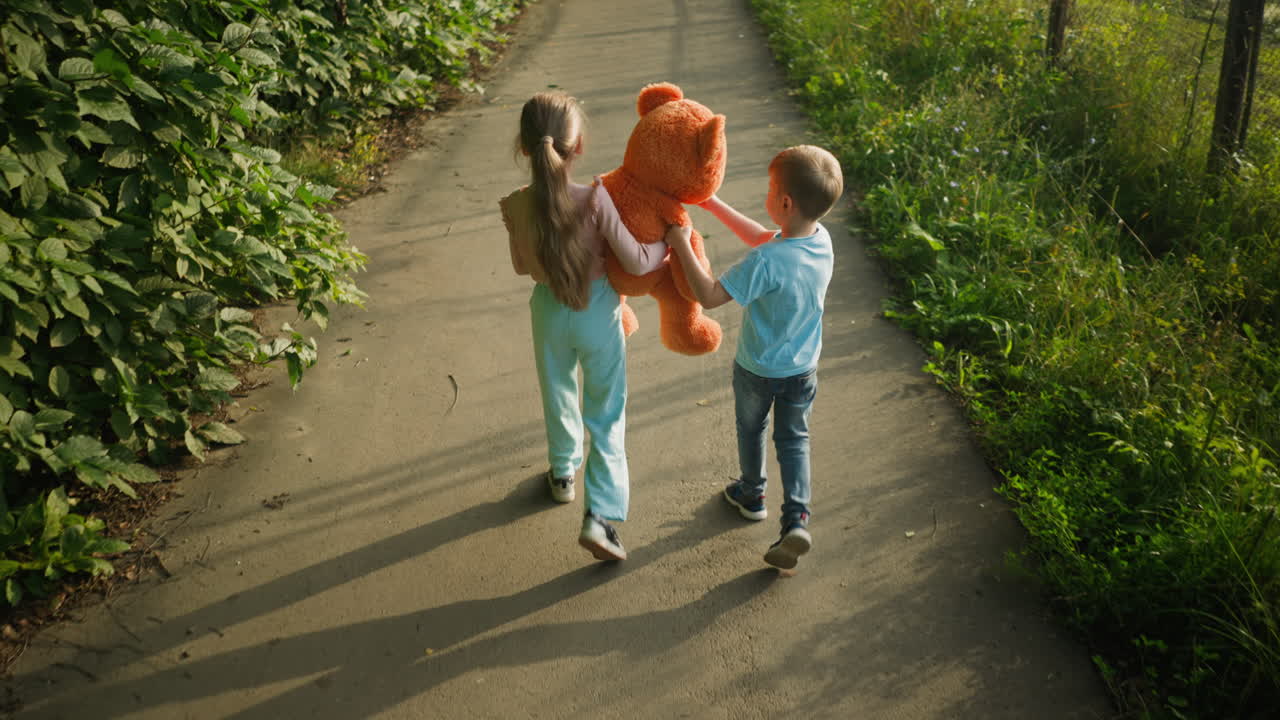 Back view of young children walking along quiet paved path bordered by green grass, with girl hugging large teddy bear and boy rubbing eyes while adjusting toy head