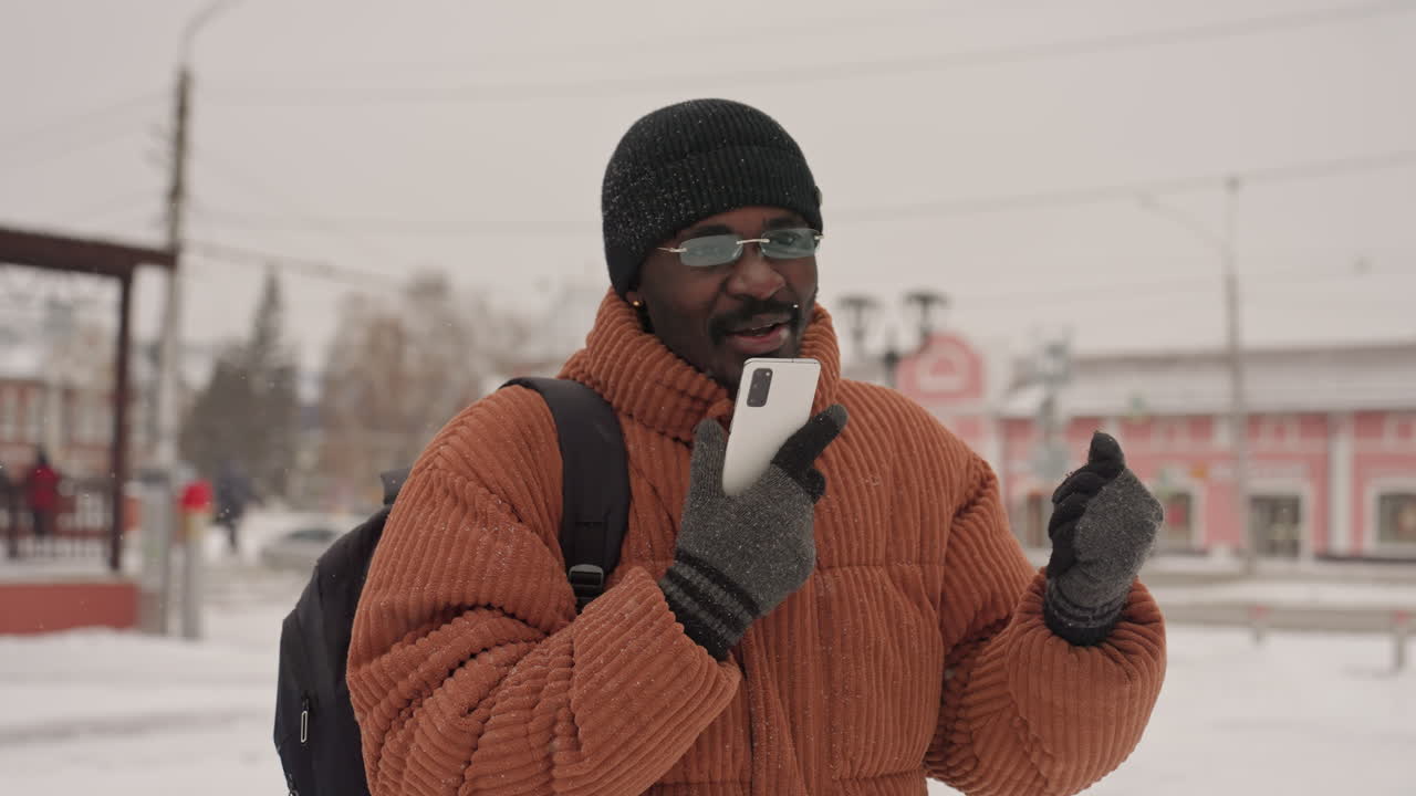 Uomo di colore in gioiosa conversazione telefonica all'aperto, con cappotto arancione spesso e zaino, tiene lo smartphone per una videochiamata, gesti animati e risate, pollice in su giocoso e reazioni espressive, città invernale.