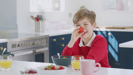 laughing boy wearing school uniform in kitchen putting strawberry on nose as he eats breakfast