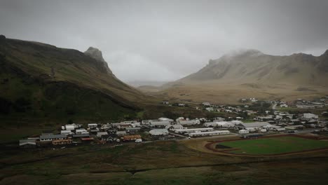 Aerial-town-vik,-houses-and-green-grassy-mountains-on-moody-foggy-day,-Iceland