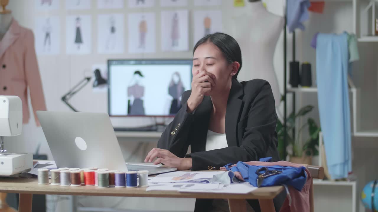 비즈니스 슈트를 입은 아시아 여성 디자이너가 노트북을 사용하며, 스튜디오에서 옷을 디자인하면서 고 잠을 자고 있습니다.