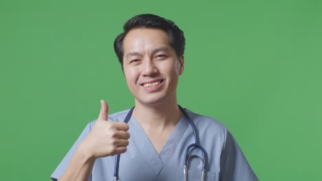 스테토스코프를 들고 있는 아시아 남성 의사의 근접 사진. 병원의 녹색 스크린 배경에서 엄지손가락을 올려놓고 미소 짓는 모습.