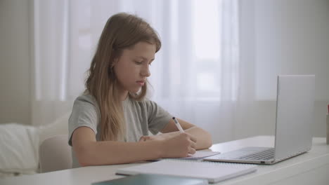 teen girl is learning distantly at home looking at screen of laptop and writing in exercise book