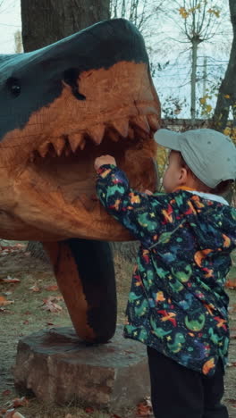lovely caucasian boy standing near the wooden sculpture of a shark. kid takes an acorn and shows it to the camera. vertical video