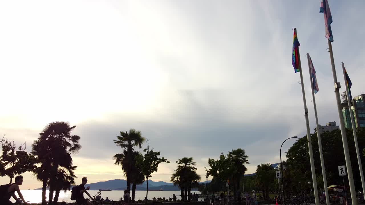 LGBTQ Flag, Pride Flag - Canadian Flag at a tropical beach where Physical Distancing is respected with people walking at a beach with palm trees during a summer sunset. 1-2