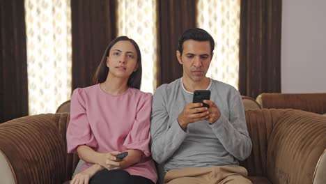 Happy-Indian-wife-watching-TV-and-husband-is-busy-in-phone