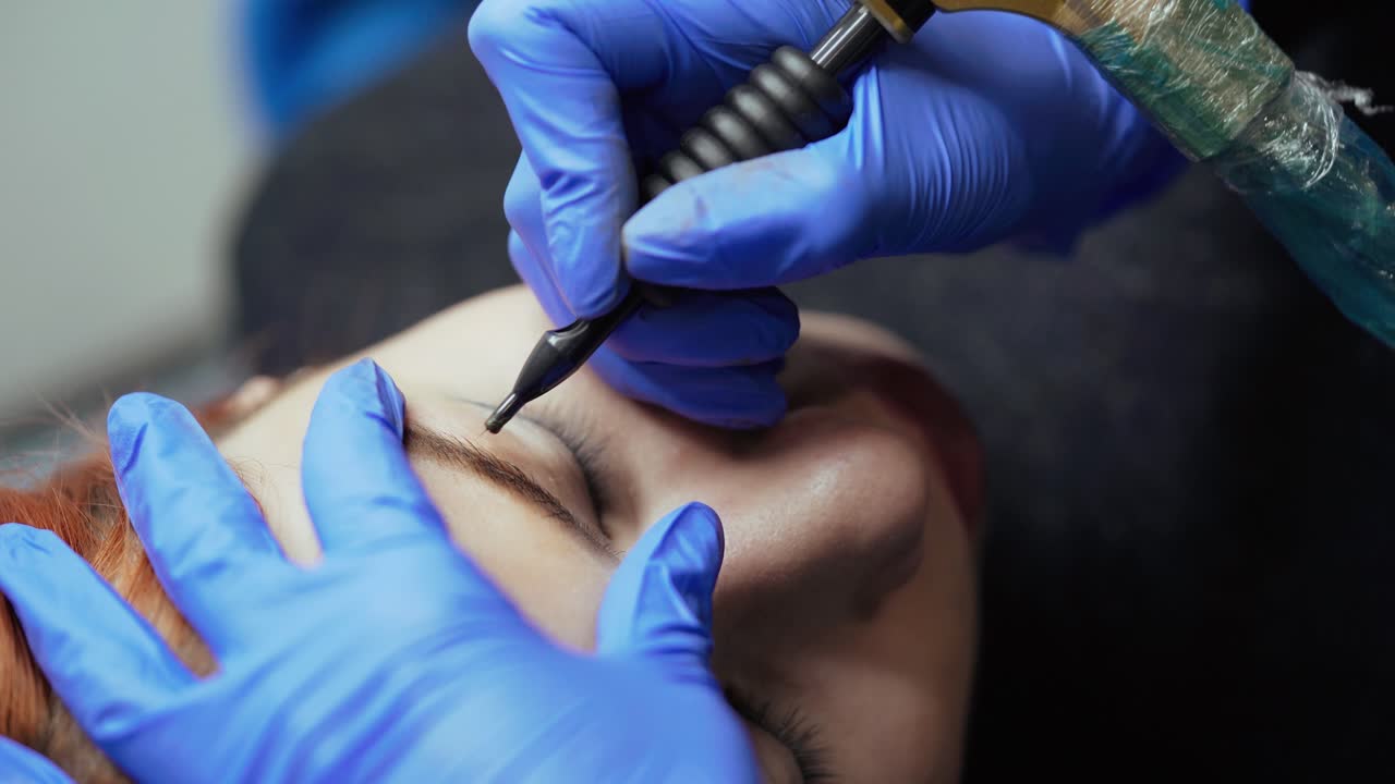 A specialist makes eyebrow tattoo with using a professional machine of permanent makeup in a beauty studio. Close-up