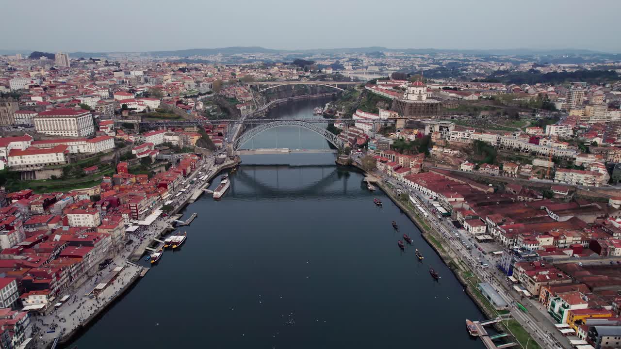 Porto cityscape with its historic buildings, traditional red tiled roofs, and winding streets lining both banks of the Douro River, featuring the iconic Dom Luis I bridge spanning across, slow aerial
