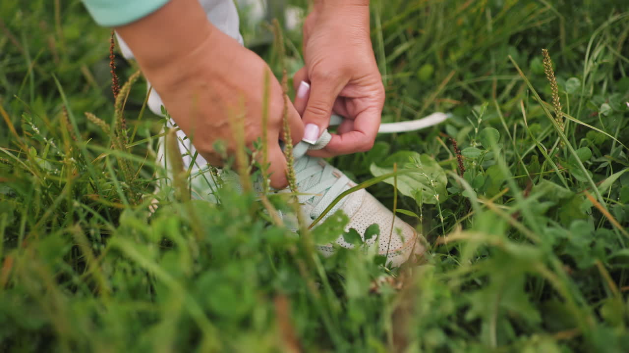 Sport woman wearing ash trouser with neatly painted short nails tying shoelaces on grassy field outdoor in summer nature preparing for workout fitness run exercise with focus on hands and shoes