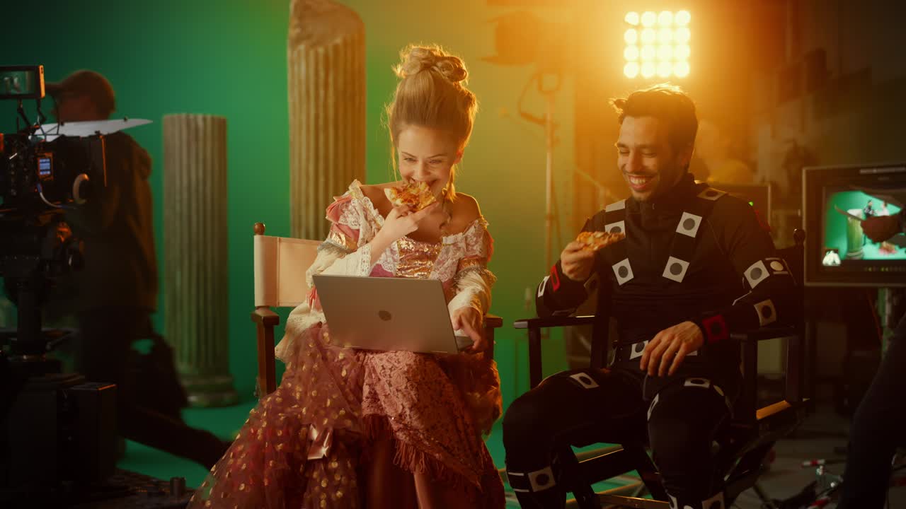 Beautiful Smiling Actress Wearing Renaissance Dress and Actor Wearing Motion Capture Suit having Lunch Break, Sitting on Chairs, Use Laptop and Talk. Studio High Budget Movie. Costume Drama Film Set