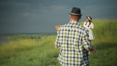 a woman wearing a white dress and hat stands in front of a canvas in a grassy field, striking a pose as a man in a checkered shirt approaches her. the scene is set in a serene outdoor environment