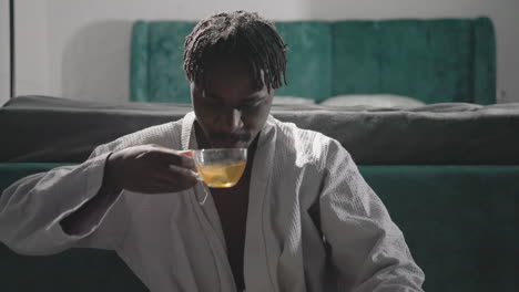 man in white robe holding tea cup, sipping tea while sitting in peaceful bedroom. soft light pours in from window, creating a calm, tranquil atmosphere perfect for a relaxing break in modern setting