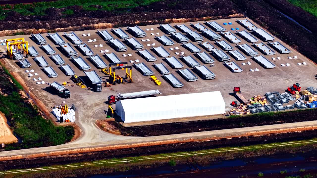 Aerial view of a storage yard filled with wind turbine components, lined in organized rows with cranes, trucks, and equipment scattered around the industrial site