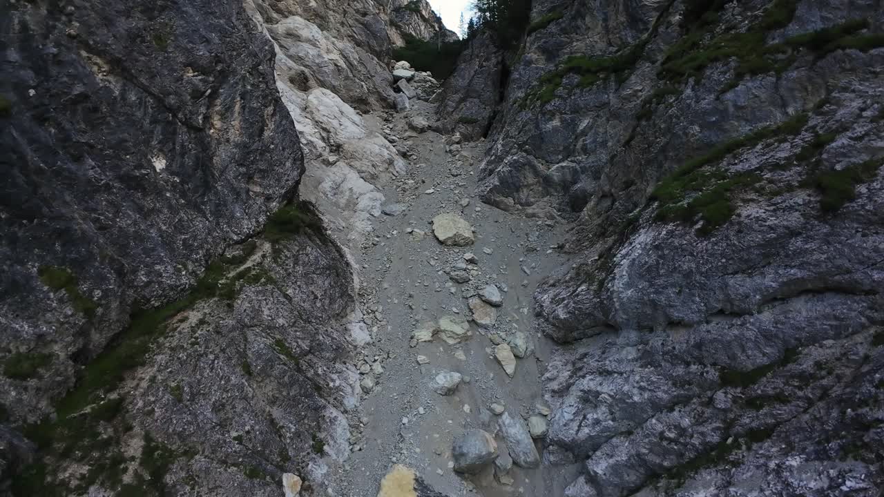 Narrow Rocky Chute Between Cliffs, Loose Stones And Compacted Scree Underfoot, Steep Vertical Walls With Moss And Exposed Roots, Tight Drone Descent Highlighting Texture And Scale, Rugged