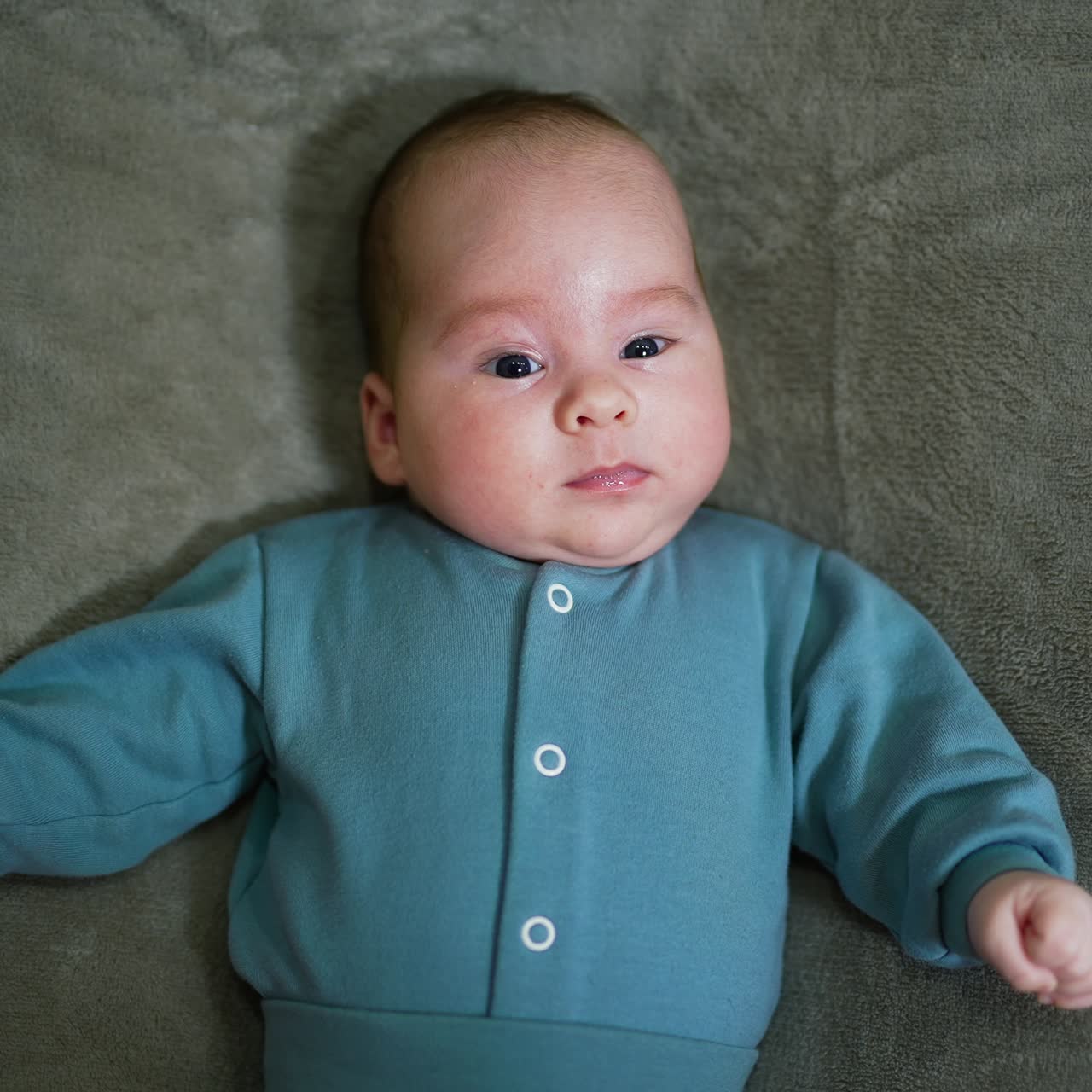 Bayi menggemaskan berbaring di atas selimut dan melihat ke kamera. Bayi laki-laki yang tenang mengenakan setelan biru dengan latar belakang abu-abu. Tampilan dari atas
