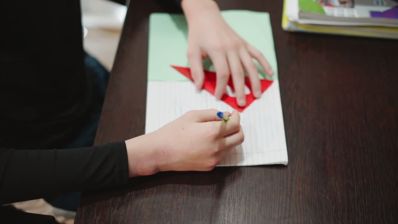 Overhead view grandson drawing geometric heart, red triangular stencil on notebook, steady pen strokes, bright daylight, study materials nearby, playful learning mood with grandma gift in mind