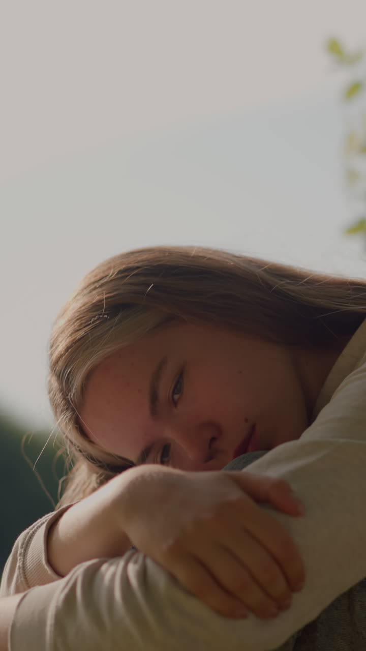 Lady sits on grassy ground, arms folded over her knees, head resting on her hand, appearing deep in thought with sunlight softly illuminating her, the blurred background includes greenery