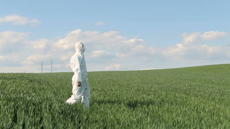 vista lateral del investigador caucásico con traje protector blanco y gafas caminando en el campo verde mientras realiza el control de plagas