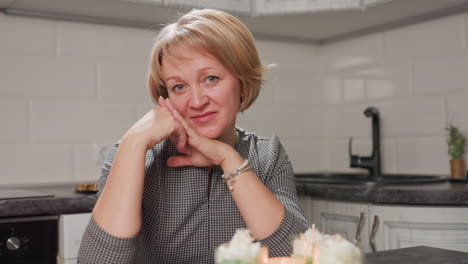 graceful lady sits in cozy modern kitchen lifting both hands warmly to cheek with soft smile, surrounded by homemade candle jars on table, reflecting calmness, satisfaction, and creative contentment