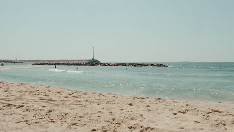 The-shore-of-israel's-blue-Mediterranean-coast-when-the-waves-hit-the-distant-breakwater,-people-in-the-water-and-a-long-antenna-can-be-seen-in-the-view