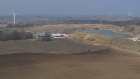 Los-Drones-Vuelan-Sobre-Un-Campo-Donde-El-Tractor-Está-Arando-Y-Los-Molinos-De-Viento-En-El-Fondo