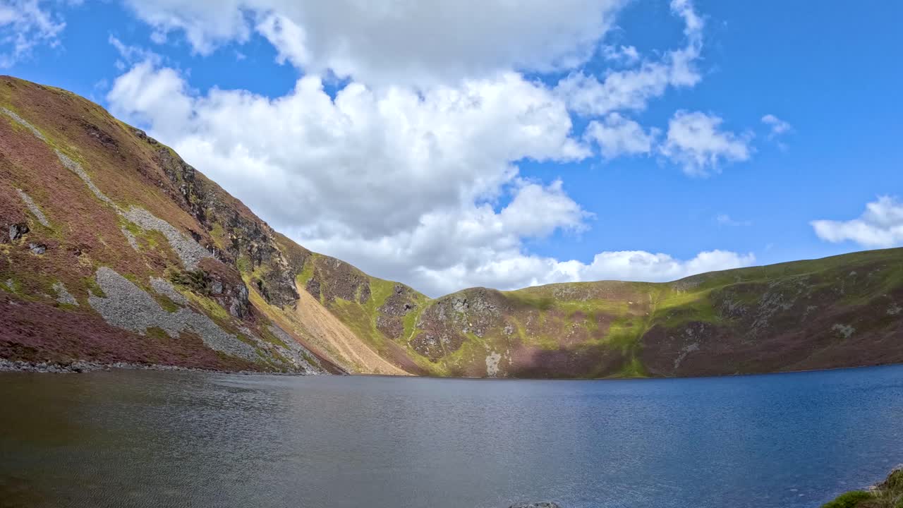 Clouds drift across a bright blue sky above Loch Brandy, surrounded by rugged hills in Glen Clova, Scotland, captured in a static wide shot