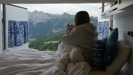 beautiful woman having cup of coffee on early morning in bed on camper van, dolomites in background