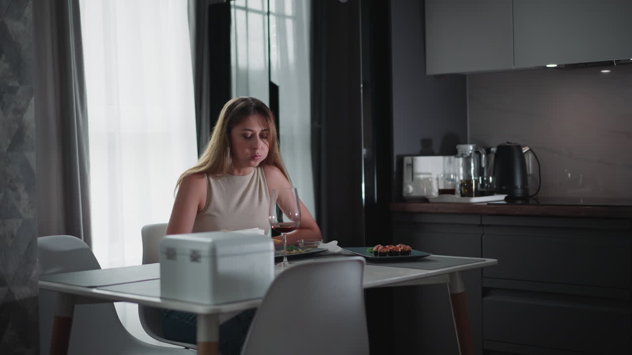 Hungry girl wearing sleeveless top sitting at modern dining table eating sushi with food in her mouth, enjoying meal quietly in cozy kitchen setting with wine glass and minimalist interior style