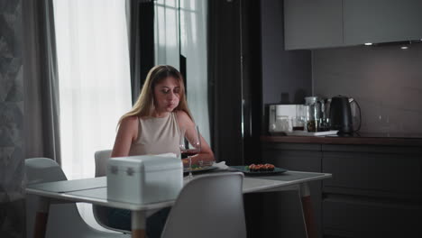 hungry girl wearing sleeveless top sitting at modern dining table eating sushi with food in her mouth, enjoying meal quietly in cozy kitchen setting with wine glass and minimalist interior style